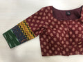 This maroon cotton blouse from VIKA Boutique, featuring a square neckline and beautiful floral patterns, includes a left sleeve with multicolored sections in green, yellow zigzags, and blue designs. The 3/4 sleeve printed blouse is displayed flat on a light wooden surface.