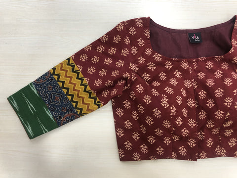 This maroon cotton blouse from VIKA Boutique, featuring a square neckline and beautiful floral patterns, includes a left sleeve with multicolored sections in green, yellow zigzags, and blue designs. The 3/4 sleeve printed blouse is displayed flat on a light wooden surface.