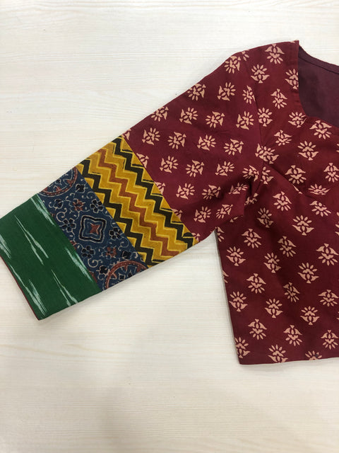 Introducing the 3/4 sleeve multicolour cotton blouse by VIKA Boutique, a maroon creation adorned with a captivating floral pattern and varied sleeve designs. It showcases vibrant green and yellow zigzags alongside an enchanting multicolored section, elegantly presented on a light wooden surface.