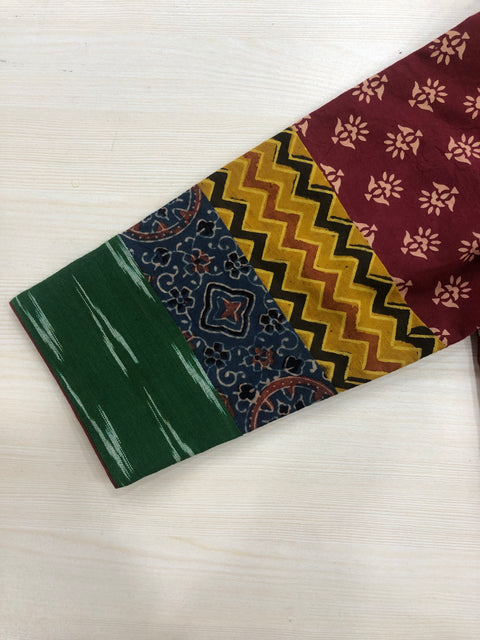 A close-up of a fabric designed for the 3/4 sleeve multicolour cotton blouse by VIKA Boutique, featuring a top section in maroon adorned with floral designs, followed by vibrant yellow zigzags, intricate geometric patterns, and a green base with white stripes, all arranged in horizontal panels.