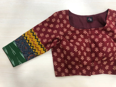 A 3/4 sleeve multicolour cotton blouse from VIKA Boutique, featuring a unique blend of beige floral patterns with green, yellow zigzag, and blue geometric prints, is beautifully displayed on a light wooden surface.